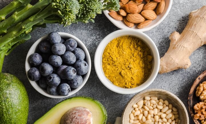 A bowl of blueberries, a bowl of spices, and a bowl of almonds surrounded by brocolli, avocado, and ginger root.