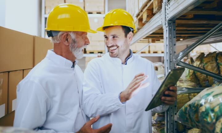 Two men weating hard hats and white coats talking in a processing plant.
